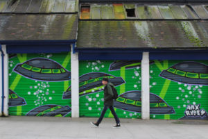 Man walking past old garages where the shutters have been painted to look like there are lots of UFOs