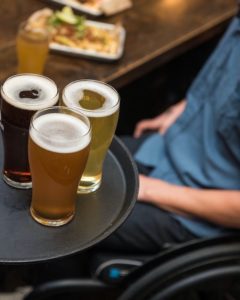 A tray with three types of beer in the foreground. In the background there is a man in a wheelchair