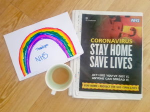 View of a newspaper advert from the government with the words Stay Home, Protect the NHS, Save Lives. Also a cup of tea and a child's drawing of a rainbow