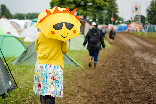 A lady in rubber boots at a muddy festival holding a yellow umbrella that has a smiley sun face